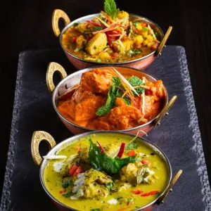 Three colorful Indian curries served in copper bowls with fresh herbs and vegetable garnishes