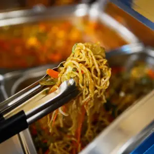 Stir-fried noodles with vegetables being served from a catering buffet tray with metal tongs